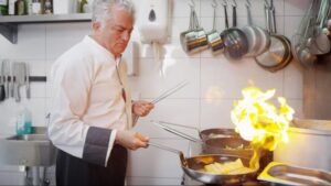 een man met grijs haar die aan het koken is met een grote flam dit is voor de Vacature horeca medewerker